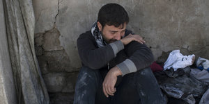 Ein Mann sitzt mit angezogenen Knieen in Trümmern vor einer Wand, er weint