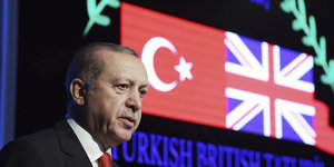 Der türkische Präsident Erdogan steht vor einem Rednerpult, hinter ihm, umkränzt von zwei digitalen Lorbeerzweigen, die türkische und die britische Flagge