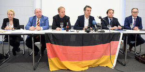 Eine Frau und fünf Männer sitzen hinter einem Tisch mit Deutschlandfahne