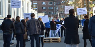 Menschen demonstrieren auf einem Betonplatz. Ein Frau hält ein Mikro, circa zwanzig Personen hören zu, einige halten Schilder