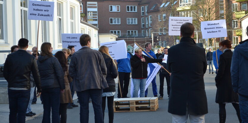 Menschen demonstrieren auf einem Betonplatz. Ein Frau hält ein Mikro, circa zwanzig Personen hören zu, einige halten Schilder