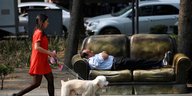 Ein Mann liegt mit verschränkten Armen auf einer Couch, die am Rande eines Bürgersteigs steht, eine Frau mit zwei Hunden eilt vorbei