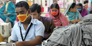 Zwei Männer und einige Frauen mit Mundschutz arbeiten an einer Nähmaschine in einer Textilfabrik in Bangladesch