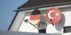 An einer Hauswand unter einem Giebel sind zwei Satellitenschüsseln installiert. Eine ist mit den Farben der deutschen Fahne bemalt, die andere mit den Farben der türkischen