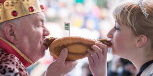 Links ein alter Mann mit Krone, rechts eine junge Frau mit Diadem. Sie beißen gemeinsam in eine Bratwurstsemmel, die in der Bildmitte ist