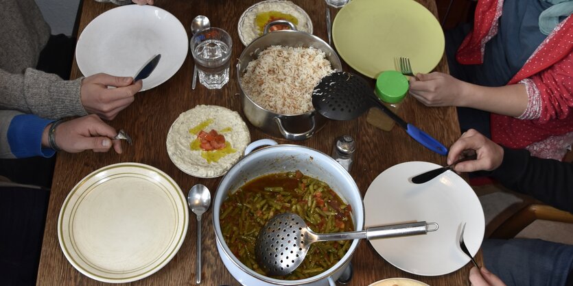 Das Essen ist fertig und steht auf dem Tisch, an dem Bilal und drei Freunde von ihm sitzen. Der Tisch, ein Holztisch, ist komplett voll. Darauf steht ein Topf mit Aldi b'set, den grünen Bohnen, ein Topf mit Reis, ein Salat und Hummus-Creme.