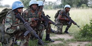 Vier Soldaten mit Gewehren und blauen Helmen hocken in Deckung am Boden