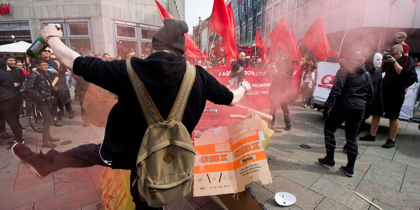 Ein Mann mit Rucksack tritt gegen einen Pappkarton, im Hintergrund viele Menschen und Rauchschwaden