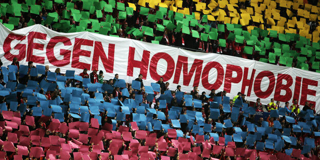 Ein großes Schild mit der Aufschrift "Gegen Homophobie", darüber und darunter halten Menschen Schilder in Regenbogenfarben hoch