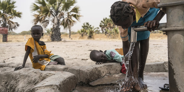 Eine Frau wäscht ihre Füße an einem Brunnen. Daneben sitzen und liegen zwei Kinder und schauen ihr zu
