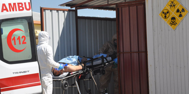 Ein Mann auf einer Liege wird von einem Helfer im Schutzanzug in einen Krankenwagen geschoben