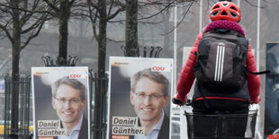 Links im Bild zwei Wahlplakate mit dem Porträtfoto eines Mannes, rechts im Bild eine Fahrradfahrerin mit Helm von hinten