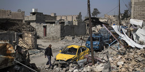 Ein Mann geht an einem Autowrack vorbei, die Häuser liegen in Trümmern