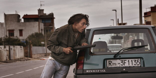 Ein junger Mann lehnt mit einer Waffe in der Hand an einem kleinen Auto