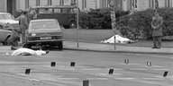 Schwarzweißfoto: Straße mit Polizeimarkierungen, stehenden Fahrzeugen und zwei zugedeckten Körpern
