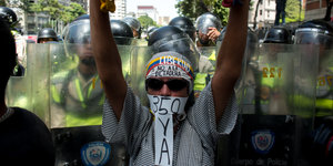 Eine Person mit Sonnenbrille und verdecktem Gesicht hält ihre Arme nach oben. Hinter ihr sind viele Polizisten oder Sicherheitsleute in gelben Westen und durschichtigen Schutzschildern