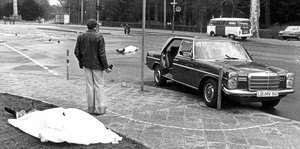 Ein Tatort: vorne links liegt ein mit einem weißen Tuch bedeckter Mensch, weiter hinten ein weiterer, vorne rechts im Bild ein Auto
