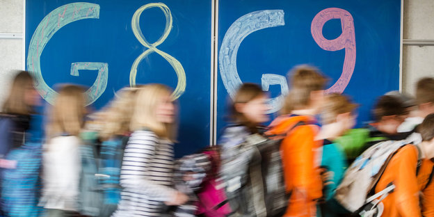 Schulkinder rennen vor einer Tafel, auf der links G8 und rechts G9 steht, nach rechts