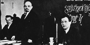 Ein schwarz-weiß Bild: Drei Männer nebeneinander, die beiden äußeren sitzen, der in der Mitte steht an einem Tisch