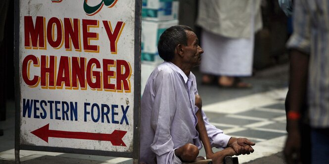 Ein gehbehinderter Mann sitzt auf dem Boden und streckt eine Hand bittend nach den Passanten aus. Direkt neben ihm steht ein Schild mit der Aufschrift "Money Changer, Western Forex"