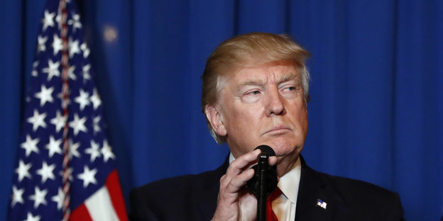 Donald Trump sitzt hinter einem Mikrofon und guckt sinnend in die Luft
