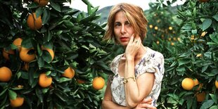 Chris Kraus steht, eine Hand an der Wange, in einem Garten zwischen Orangenbäumen