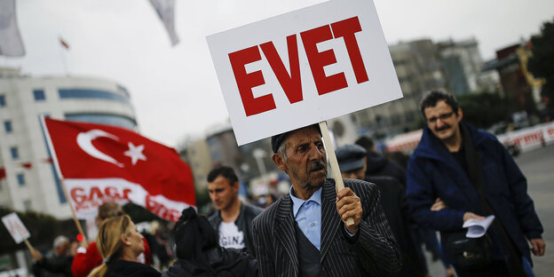Im Vordergrund ist ein älterer Mann mit einem Schild auf dem "Evet", also "Ja" steht, im Hintergrund stehen weitere Menschen, einer schwingt eine türkische Flagge