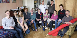 Eine Gruppe von Menschen sitzt im Zimmer einer Wohnung auf Stühlen und einem Bett