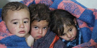 drei kleine Kinder, die, eng aneinandergeschmiegt, traurig in die Kamera blicken