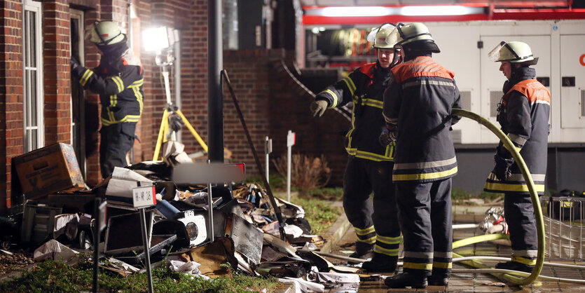 Feuerwehrleute stehen nachts mit Löschschlauch in einer Pfütze vor einem Kellerfenster.