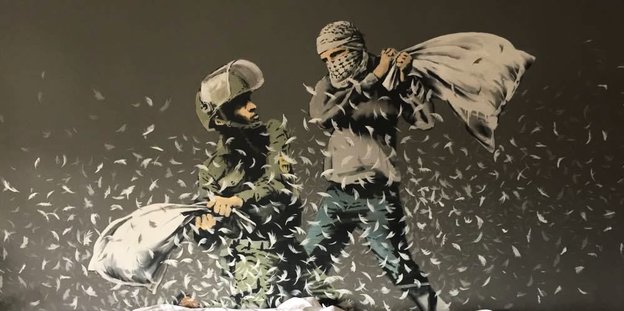 Ein vermummter palästinensischer Aktivist und ein israelischer Soldat kämpfen mit Kissen, die Federn stieben