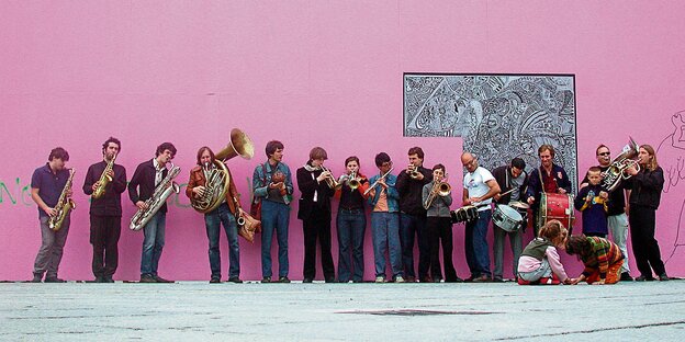 16 Musiker mit Blasinstrumenten und Schlagzeugen vor einer rosa Wand.