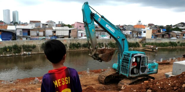 Ein kleiner Junge in einem Barcelona-Trikot schaut auf das Flussufer, auf dem ein Bagger in den Schlammassen gräbt. Im Hintergrund sieht man die Hochhäuser Jakartas.