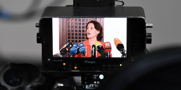 Auf einem Kamerabildschirm sieht man: Eine Frau steht vor vielen Mikrofonen