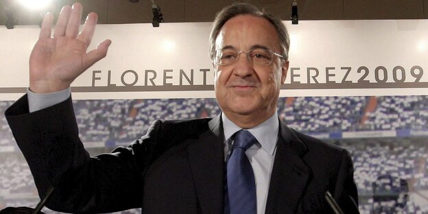 Ein lächelnder winkender Mann in Anzug und Krawatte vor den Rängen eines Stadions und dem Schriftzug Florentino Pérez.