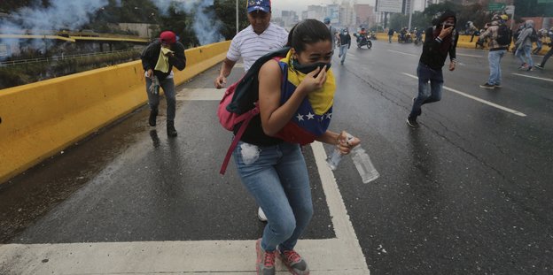 Eine Gruppe Demonstranten läuft auf einer Straße auf den Betrachter zu. Sie werden mit Tränengas beschossen. Im Vordergrund eine Frau. Sie hält zwei Flaschen in der linken Hand und bedeckt ihren Mund mit einer venezualischen Fahne.