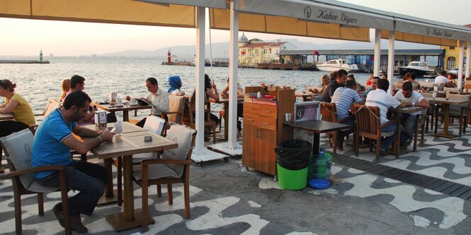 Menschen sitzen im Freien in der Abendsonne an Tischen in einem Café am Wasser.