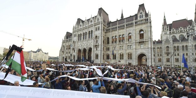 Eine Menschenmenge trägt ein langes Stück Papier über den Köpfen, im Hintergrund ein großes Gebäude
