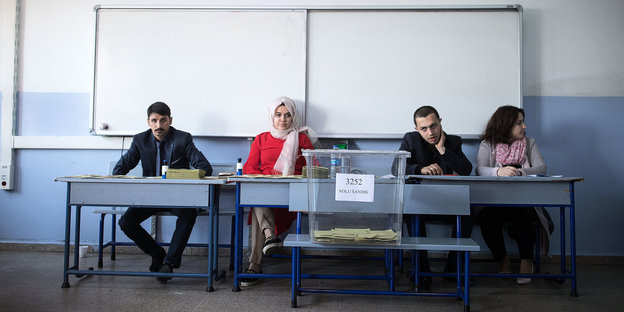 zwei Männer und zwei Frauen hinter einem Tisch, davor eine große durchsichtige Wahlurne