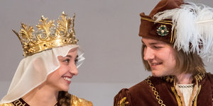 Eine Frau mit Schleier und Krone lächelt einen Mann mit Hut an