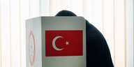 Ein Mensch sitzt in einem Wahllokal hinter einem Sichtschutz, der das Symbol der türkischen Fahne trägt