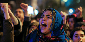 Eine junge Frau mit Kopftuch ruft in einer Menschenmenge, sie hält ein Transparent