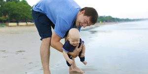 Ein Vater steht barfuß am Strand, im Arm hält er ein kleines Kind. Er beugt sich vor, um dem Kind den Sand zu zeigen und lacht
