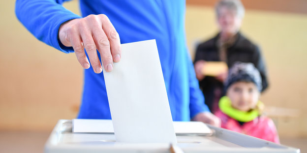 Eine Person in blauem Pulli steckt einen Wahlzettel in eine Wahlurne.