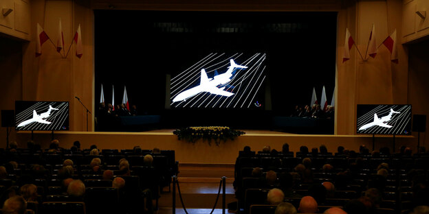 Eine Theatersaal aus der Zuschauerperspektive, auf der Bühne steht eine Leinwand, auf der ein weißes Flugzeug abgebildet ist