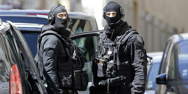 Zwei Polizisten, die Schutzwesten und Kopfbedeckungen tragen, so dass nur ihre Augen zu sehen sind, stehen zwischen Autos