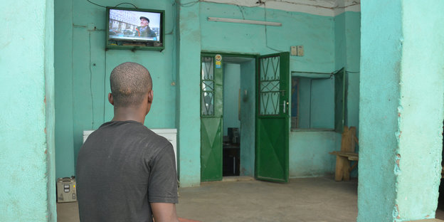 Alpha Mahamadou Diallo sitzt mit dem Rücken zum Betrachter in einem türkisfarbenen Raum und schaut zu einem Fernseher, der an der Wand hängt