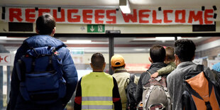 Geflüchtete vor einem Schild, auf dem auf Englisch „Willkommen Geflüchtete“ steht