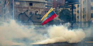 gelblicher Rauch von Tränengas zieht über eine Straße in Caracas, mittendrin halten Demonstranten die venezolanische Flagge hoch