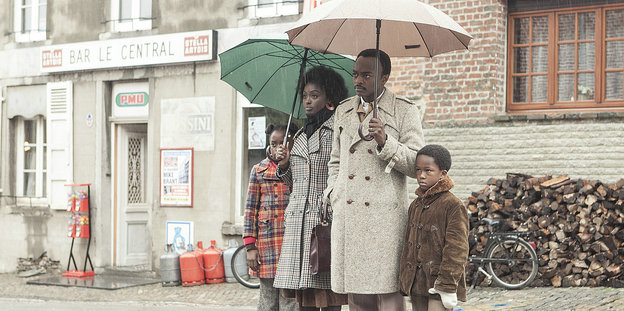 Vor einer Häuserfassade steht eine schick gekleidete, schwarze Familie. Von links nach rechts: ein Mädchen, eine Frau, ein Mann und ein kleiner Junge. Die Eltern halten Regenschirme, denn es schüttet wie aus Eimern